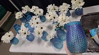 20 blue hobnail glass vases, some filled with faux white daisy flowers, displayed on a table.