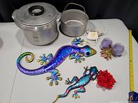 Photo showing two colorful lizard metal wall hangings, vintage aluminum stove pot with perforated insert basket and lid, purple candles, flower decor, razor, and an electric plug adapter.