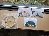 Photo showing the three embroidery design patterns laid out on a window ledge along with embroidery tools such as wooden hoop, needle, floss, and fabric piece.