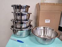 Four stainless steel nesting bowls stacked with a colander beside them on a small table, next to cardboard boxes and a blue marker pen for scale.