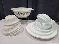 Stack of white Corelle plates and bowls with some bowls stacked to the side and a white Pyrex dish with brown floral vine pattern elevated on a clear stand.