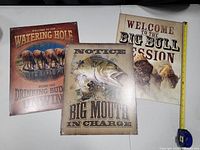 Three metal signs each 16 x 12.5 inches with fishing, horse, and bull drinking themes laid flat with tape measure for scale.