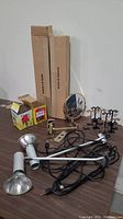 Overview of the lot showing two silver-colored adjustable wall mount lights with bulbs in boxes, cords and plugs, a round brass framed mirror on a stand, and several earring display stands in black and beige.