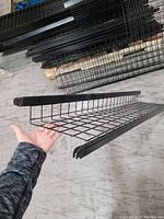 Photo of a hand holding a black metal wire basket shelf showing the wire grid base and raised edges for storage