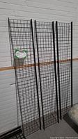 Three black metal wire mesh upright panels leaning against wall showing height and grid detail.