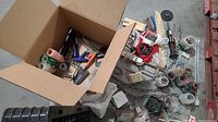 Box filled with various tools including a hand saw, chains, rollers, and assorted hardware items spilled on the floor and beside the box.