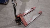 Side view of the red pallet jack on a concrete floor showing handle and forks.