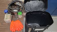 Open black hard plastic rolling case with various tools and accessories spread next to it on a table including bright orange gloves, a coiled hose, containers of nails and hardware.