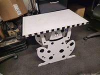 Front view of handmade wooden table showing a white cat figure with black spots holding up the rectangular tabletop with black and white striped edges.