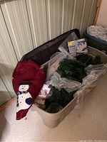 Large plastic storage tote partially open showing a red Christmas tree skirt with a snowman design and some artificial green garland inside, tote lid leaning behind