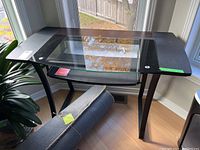 Photo of a computer desk with a tinted tempered glass top, black frame, and curved black metal legs. A pull-out keyboard tray is underneath the glass surface.