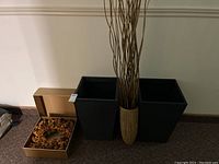Two black woven patio planters, decorative beige vase with tall reeds, and a festive wreath in gold box visible.