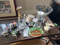 Wide shot showing all items on table including Royal Doulton figurine, glass carafe with cup, tea cups, elephant salt and pepper shakers, decorative plate, white ceramic birds, and glass vase