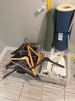 Photo showing tote with mixed wooden clothes hangers, a green rolled bath mat, an off white rolled rug secured with strap, and other mats inside clear bins.
