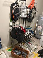 View of Household lot showing 4 backpacks on wall hooks, orange reusable bags, black metal rack with shovels and rakes, clear plastic storage bins with small hardware and light bulbs near water heater.