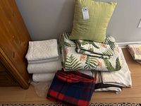 Lot of assorted linens arranged including green quilted throw pillow on top of floral comforter, red navy plaid fleece blanket, multiple folded white and beige pillow cases or sheets.