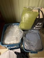 Two blue plastic totes filled with assorted linens including towels, sheets, and pillows covered in plastic bags along with larger pillows and bags resting on top.