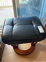 Black vinyl cushioned ottoman with visible tag and price sticker on top, sitting on wood floor near window light.