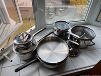 Overview of assorted stainless steel pots and pans on window sill, including frying pans, saucepans with lids and mixing bowls