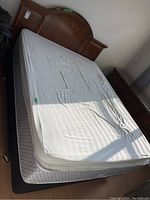 Full view of queen bed with mattress and boxspring, white sheet covers mattress and wooden headboard visible.