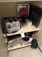 The cabinet shelf displays kitchen items including two toasters, aluminum foil trays, cooling racks, frying pans, a small crockpot, a small food processor, and oven mitts.