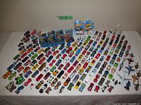 Wide shot of full set of approximately 120 diecast toy cars spread out on table, including loose cars and packaged items. Various colors and models, some motorcycles, trucks, cars, airplanes.