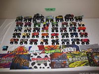 Full view of 40 Monster Jam toy trucks arranged in rows on a table along with 8 colorful Monster Jam-themed fabric flags displayed in front.