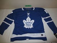 Front view of blue Toronto Maple Leafs hockey jersey with white maple leaf emblem and lace-up neck detail, size and branding tag on hem visible.