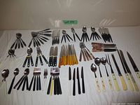 Overview of all vintage cutlery laid out on a white cloth showing multiple sets with various plastic handle colors including black, yellow, cream, and tortoiseshell.