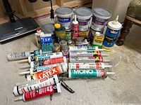 Photo showing various tubes and containers of adhesives, silicones, latex, lubricants, and other related products arranged on a floor.