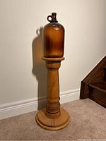 Full view of the amber glass jug placed atop the wooden pedestal against a wall and carpeted floor.