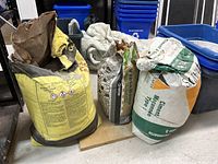Three bags including two opened bags of St Marys masonry cement and one opened bag of VersaBond thin-set mortar stored indoors. Bags are dry and loose with visible wear from opening.