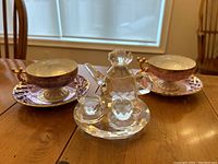 Two matching Japanese tea cups and saucers in pink-purple with gold accents, one cup showing a chip on the rim, alongside a Sorelle crystal decorative miniature tea set on a wooden table.