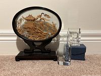 Frontal view showing the carved wood artwork behind oval glass frame with black wooden stand plus two clear crystal figurines on the right side.