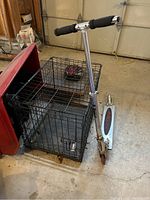 Photo showing black metal folding dog crate next to silver kick scooter on garage floor.