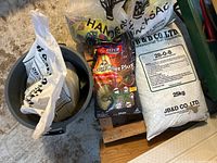 View of two white plastic bags of JB & D Co. Ltd. fertilizer and red/black packaging of Bayer Advanced Grub Killer Plus in a gray bucket and nearby on the floor.