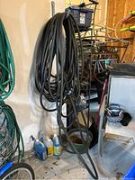 Two large black garden hoses hanging coiled on a wall hook in a garage, alongside other items like cleaning solutions and a bicycle rack.