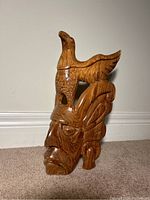Full view of the wooden sculpture showing human face with a bird on top, placed on carpeted floor against baseboard.