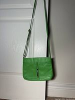 Front view of small green leather crossbody bag with vertical zipper pocket and strap hanging on door handle.