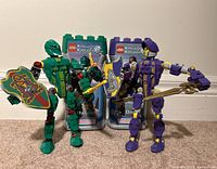 Two assembled Lego Knights Kingdom figures with their original packaging and game cards in the background. Rascus figure is green, Danju figure is purple. Both hold shields and weapons.