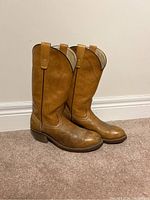 Pair of tan leather cowboy boots with stitching patterns and pull tabs, standing upright on carpet against a white wall.