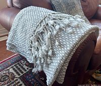 Light grey woven bohemian style blanket with white textured dot pattern and twisted tassels on the edges, draped over a brown leather sofa.