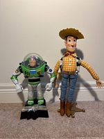 Photo showing full body views of Buzz Lightyear and Woody figures standing upright with Woody's stand visible.