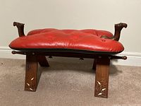 Side view showing carved camel heads on stool armrests, red leather cushion, and wooden legs with decorative inlays.