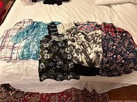 Eleven blouses and shirts laid out on a bed showing various colors and patterns including plaid, tie-dye, floral, and abstract designs
