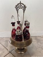 Front view of five ruby etched glass bottles with two clear stoppers and three silver tops in silver-plated tray with ornamental handle.