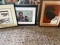 Three framed artworks side by side on carpeted floor: abstract canoe print (left), color photograph (center), geometric brown abstract (right)