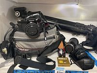 Full view of Nikon Nikkormat camera on Gamrac carry bag, black tripod behind, and various lenses and straps alongside on white surface.