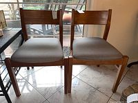 Pair of mid-century modern style dining chairs with wood frames and grey upholstered seats, positioned side by side on a tiled floor in front of a window during daytime.