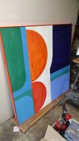 Front angled view showing the full painting and colorful geometric shapes in orange, blue, green, white, and purple in an orange aluminum frame.